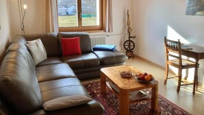 Ett mysigt vardagsrum med en lädersoffa, ett träbord med frukt på, en trästol och ett fönster med utsikt över hus.