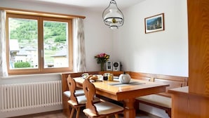 En matplats med ett träbord och stolar, ett element, ett fönster med gardiner, en vägglampa och en inramad tavla.