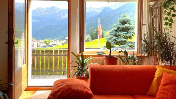 Una habitación con sofá, plantas en macetas y vistas a montañas e iglesia.