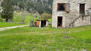 A rustic stone building with a wooden door and window, surrounded by a lush green landscape and a mountain backdrop.