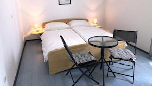 A single bed with a wooden headboard, two bedside lamps, a small round table, two chairs, and a framed picture on the wall. - The Blue Line Ferienwohnung mit Balkon by Interhome (Rab/Kampor)