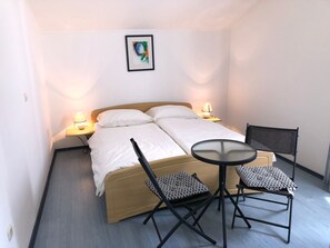 Un lit simple avec une tête de lit en bois, deux lampes de chevet, une petite table ronde, deux chaises et un tableau encadré accroché au mur.