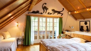 Une chambre confortable située sous les toits, avec des poutres apparentes, une grande fenêtre ornée de rideaux verts et une décoration murale représentant un chat.