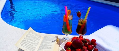 A poolside setting with a book, two drinks with straws, and a bowl of fruit.