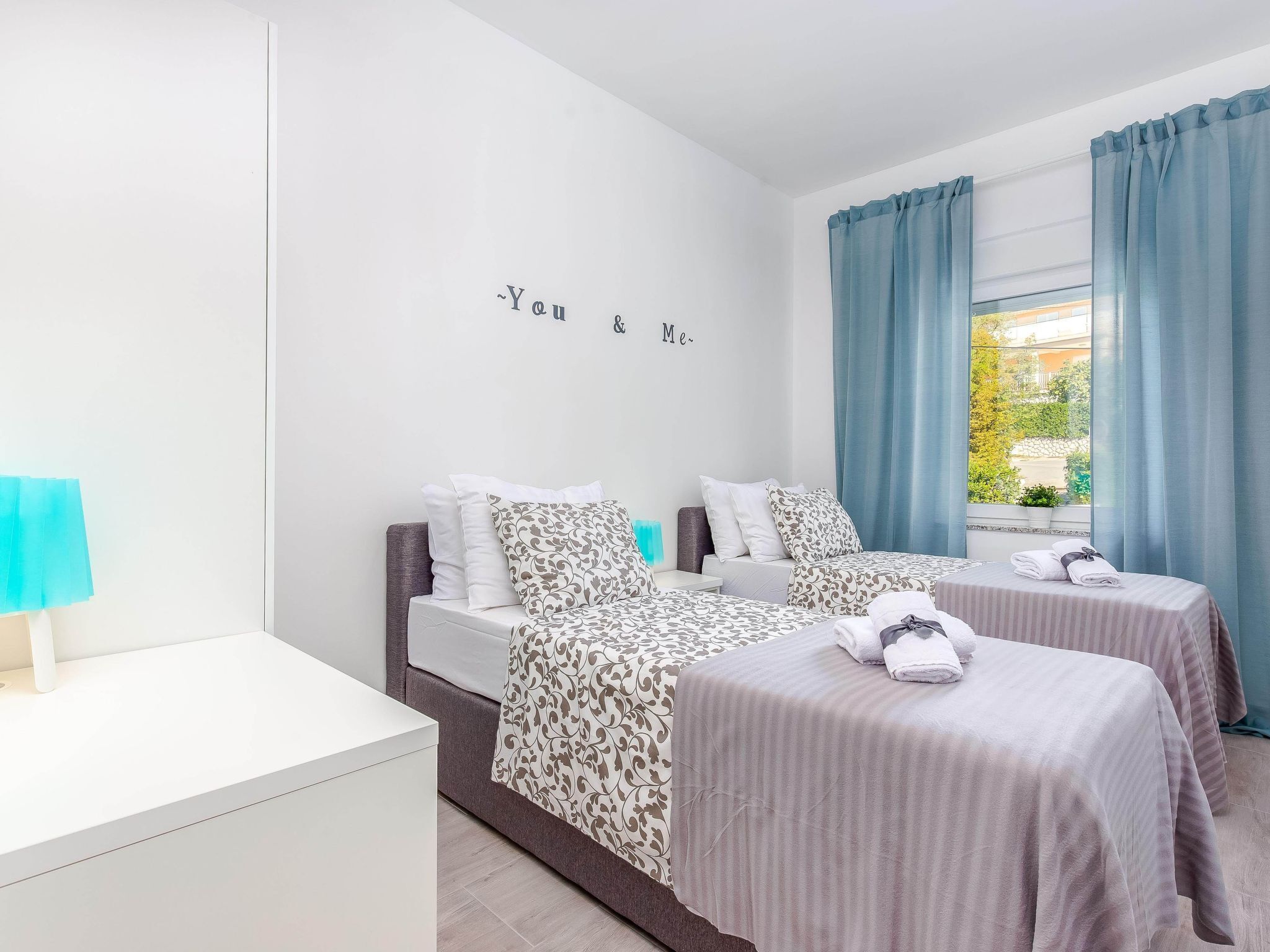 A hotel room with two beds, a white nightstand, and a window with blue curtains.