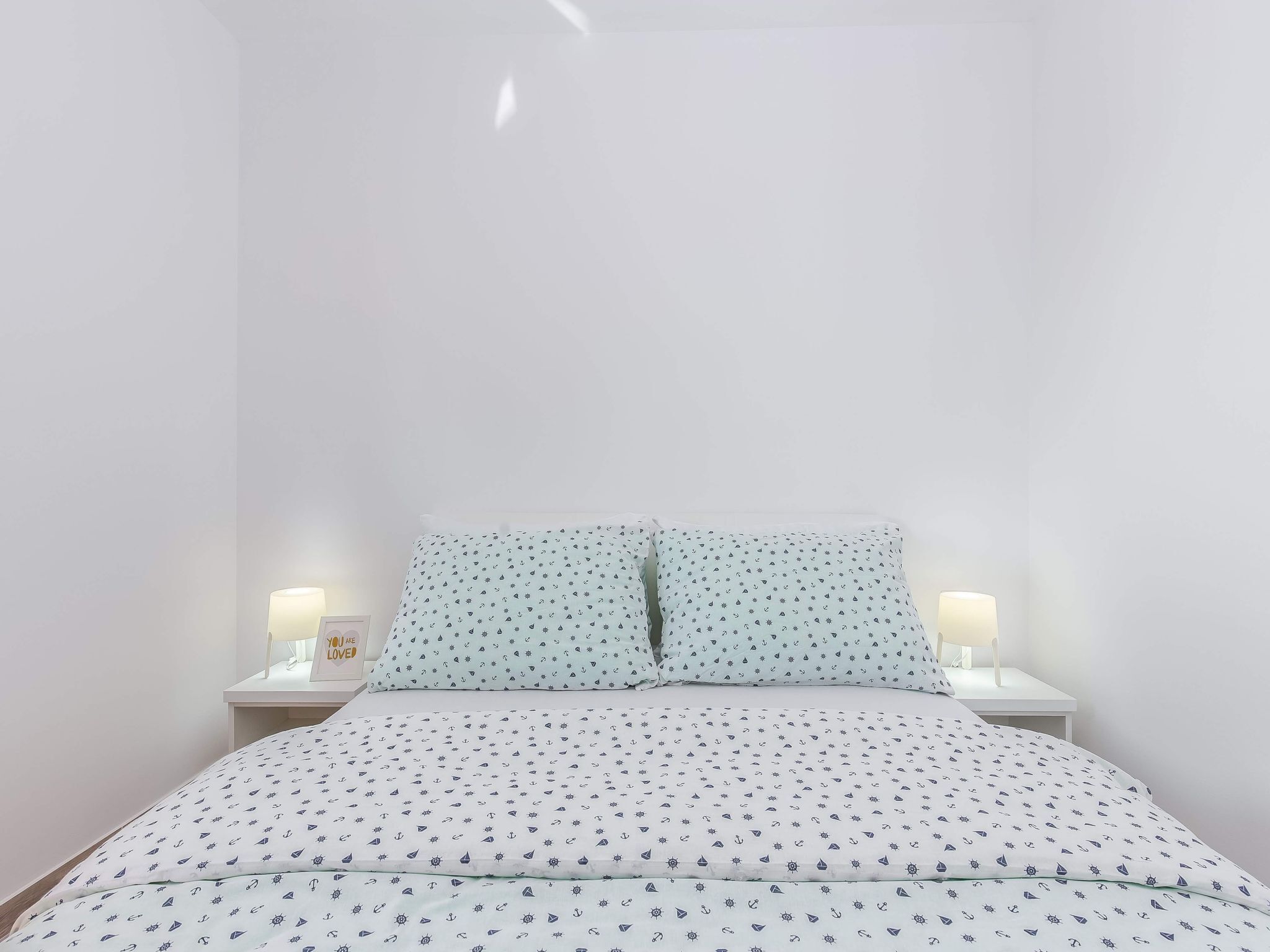 Una cama bien hecha con una colcha blanca y azul claro estampada, dos almohadas a juego y dos mesitas de noche con lámparas.