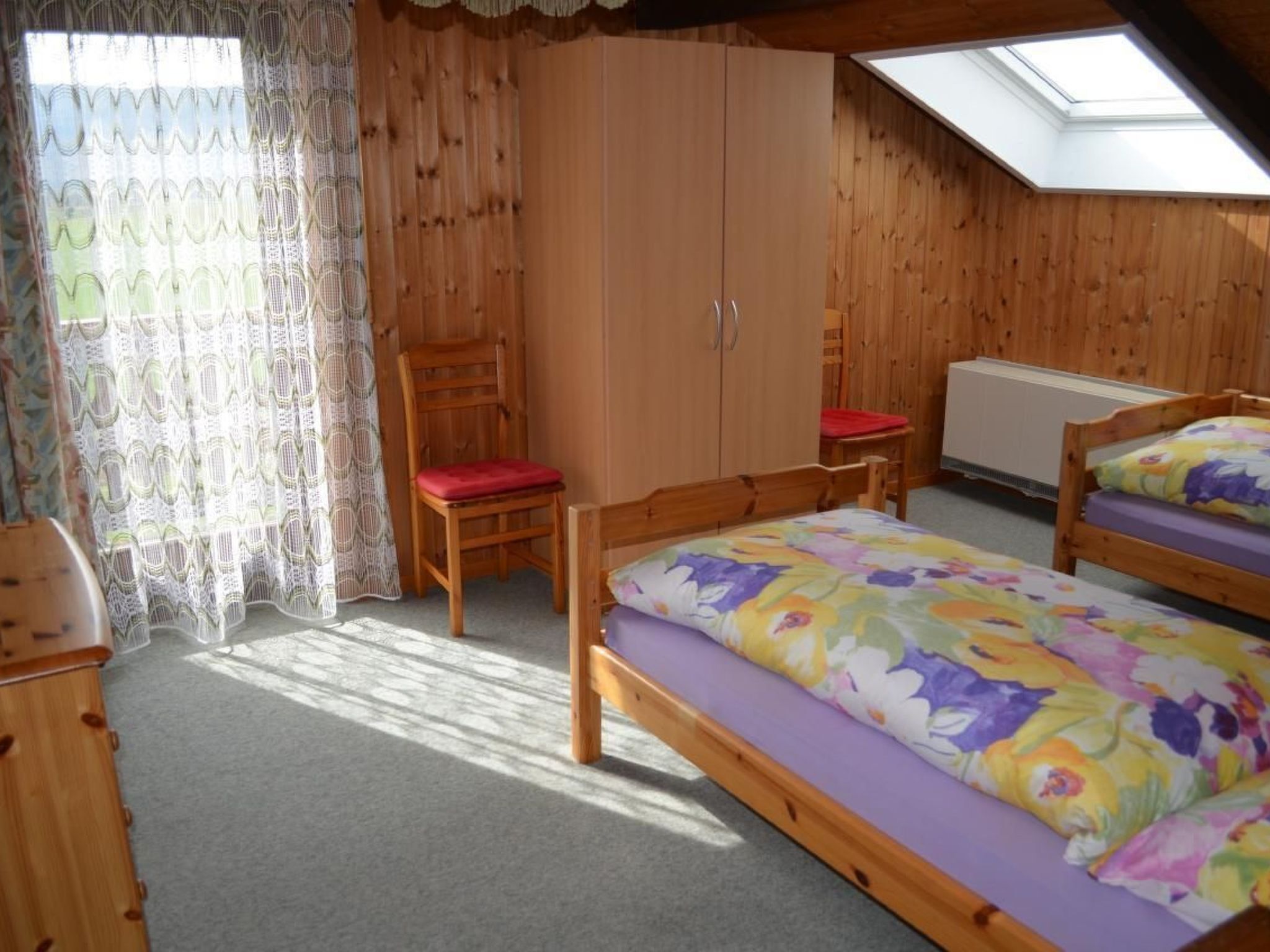A cozy room with wooden walls, a skylight, two beds with floral bedding, a chair with a red cushion, and a wardrobe.