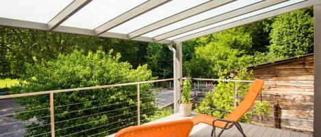 Ein überdachter Außenbereich mit zwei orangefarbenen Stühlen, einem Holzdeck und Blick auf einen üppigen grünen Garten.