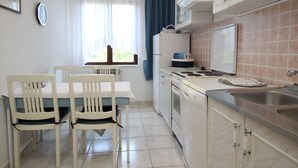 A compact kitchen with white cabinets, a dining table with chairs, a built-in oven, and a large window with curtains. - Apartment Andjela by Interhome (Krk/Šilo)