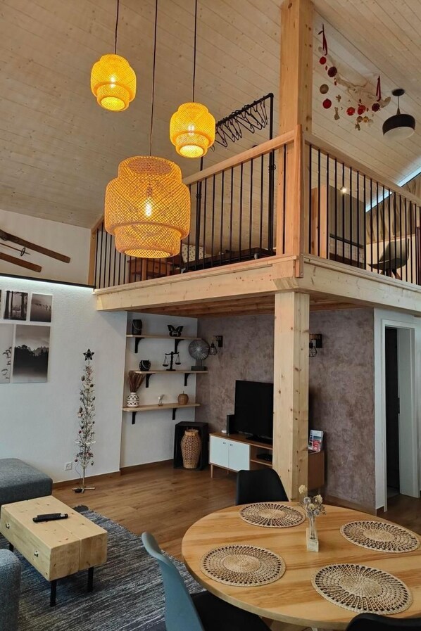 A spacious living area with a wooden ceiling, a balcony with a railing, and a dining table with woven placemats.