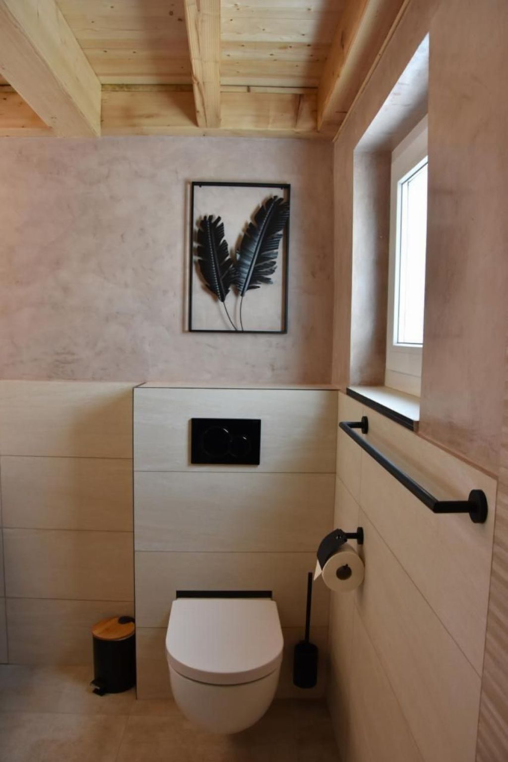 Un bagno piccolo con un water bianco, un cestino nero, un portasciugamani per carta igienica fissato alla parete e un'opera d'arte con piume in cornice appesa al muro.
