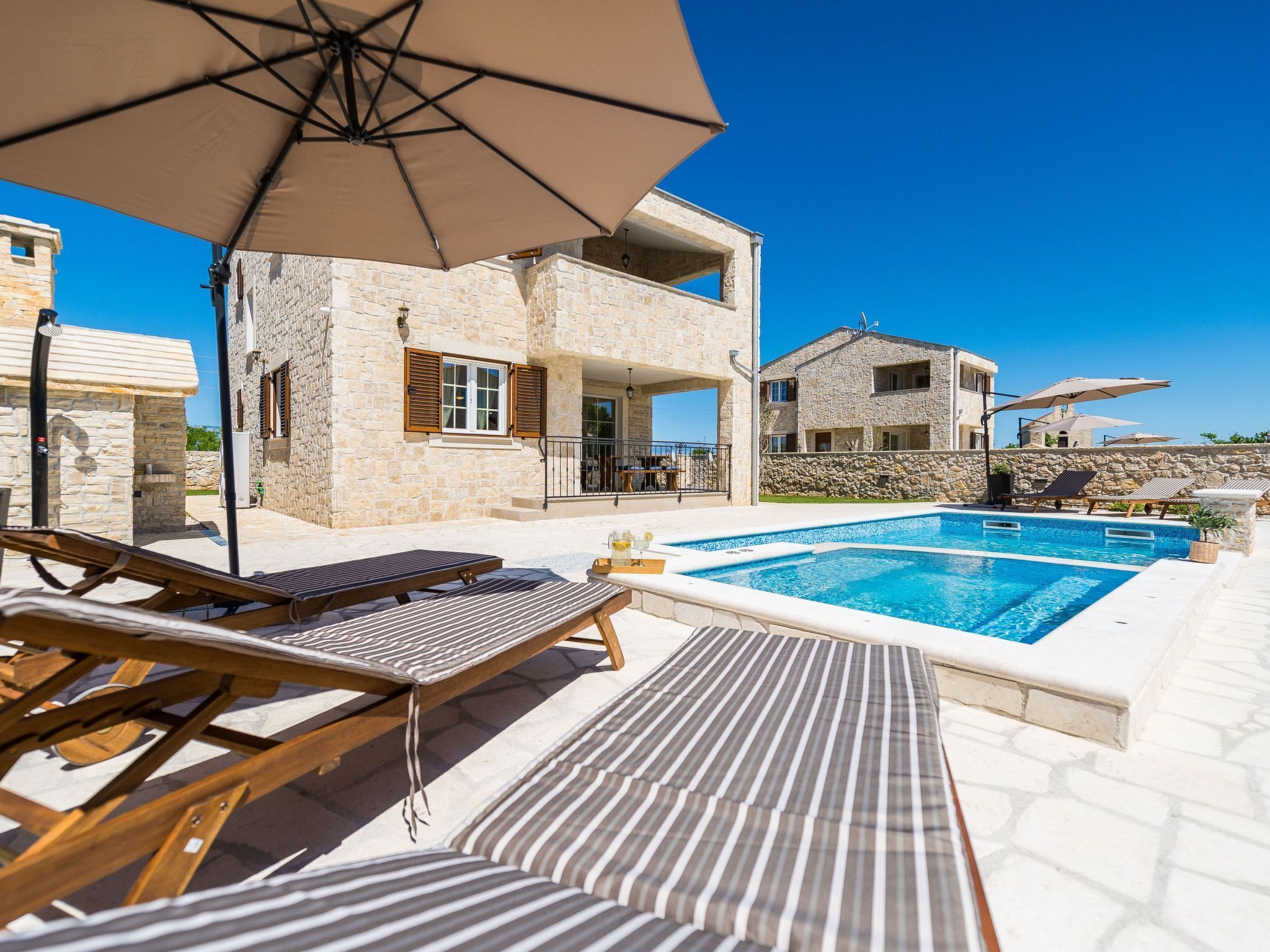 Ein Poolbereich mit Liegestühlen und Sonnenschirmen, ein Steinhaus mit Balkon und ein klarer, blauer Himmel.