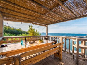 Eine Holzterrasse im Freien mit einer Bank und einem Tisch, mit Blick auf ein Gewässer und viel Grün.