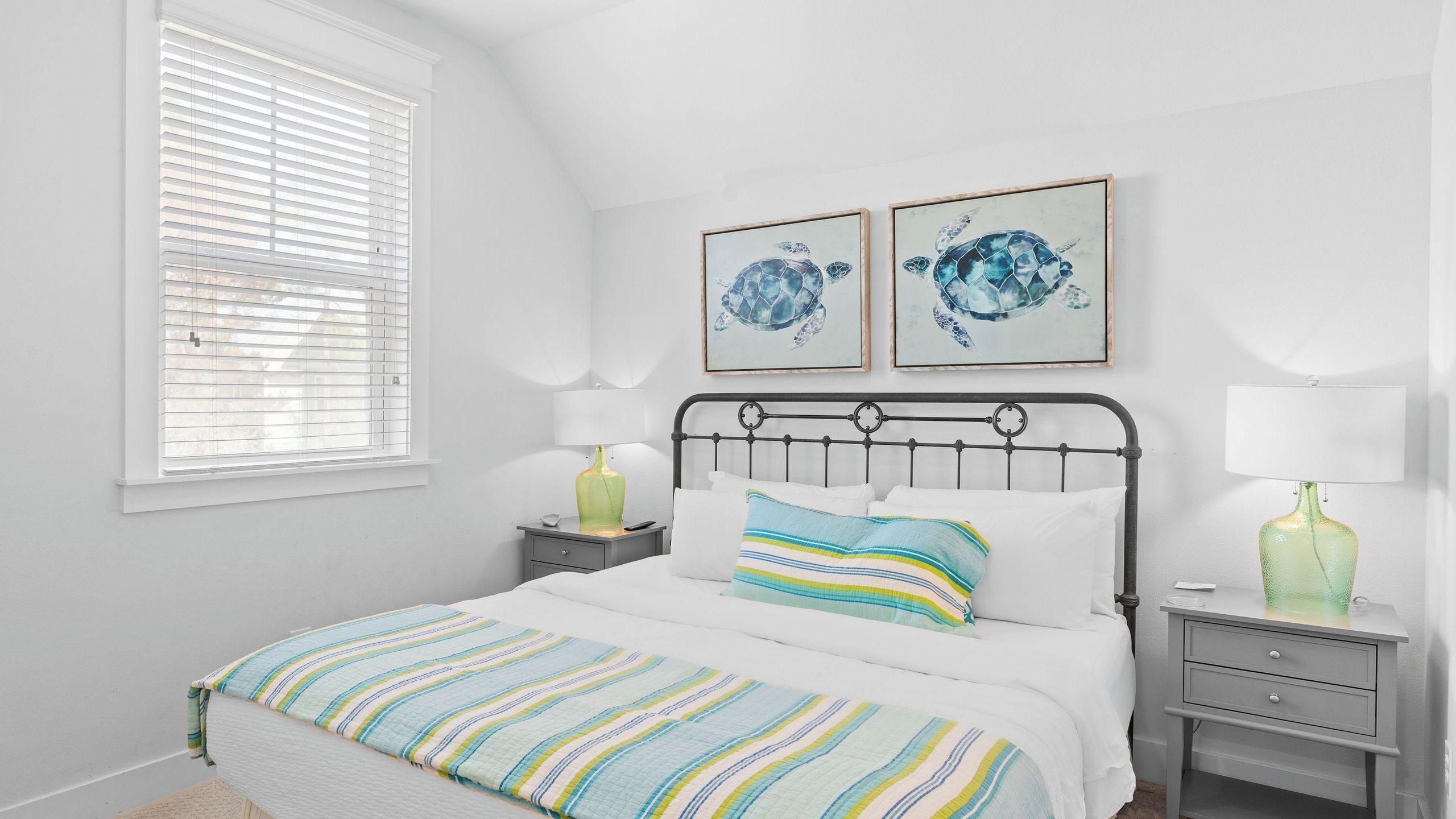 A bedroom with a bed, a nightstand, a lamp, a window with blinds, and two framed pictures on the wall.