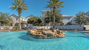 A clear swimming pool with a central rock feature, surrounded by lounge chairs and palm trees.