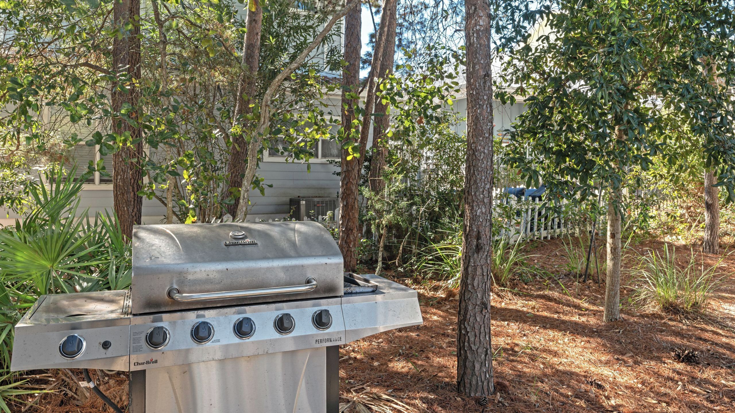A Char-Broil gas grill in a wooded backyard with trees and a white fence in the background.