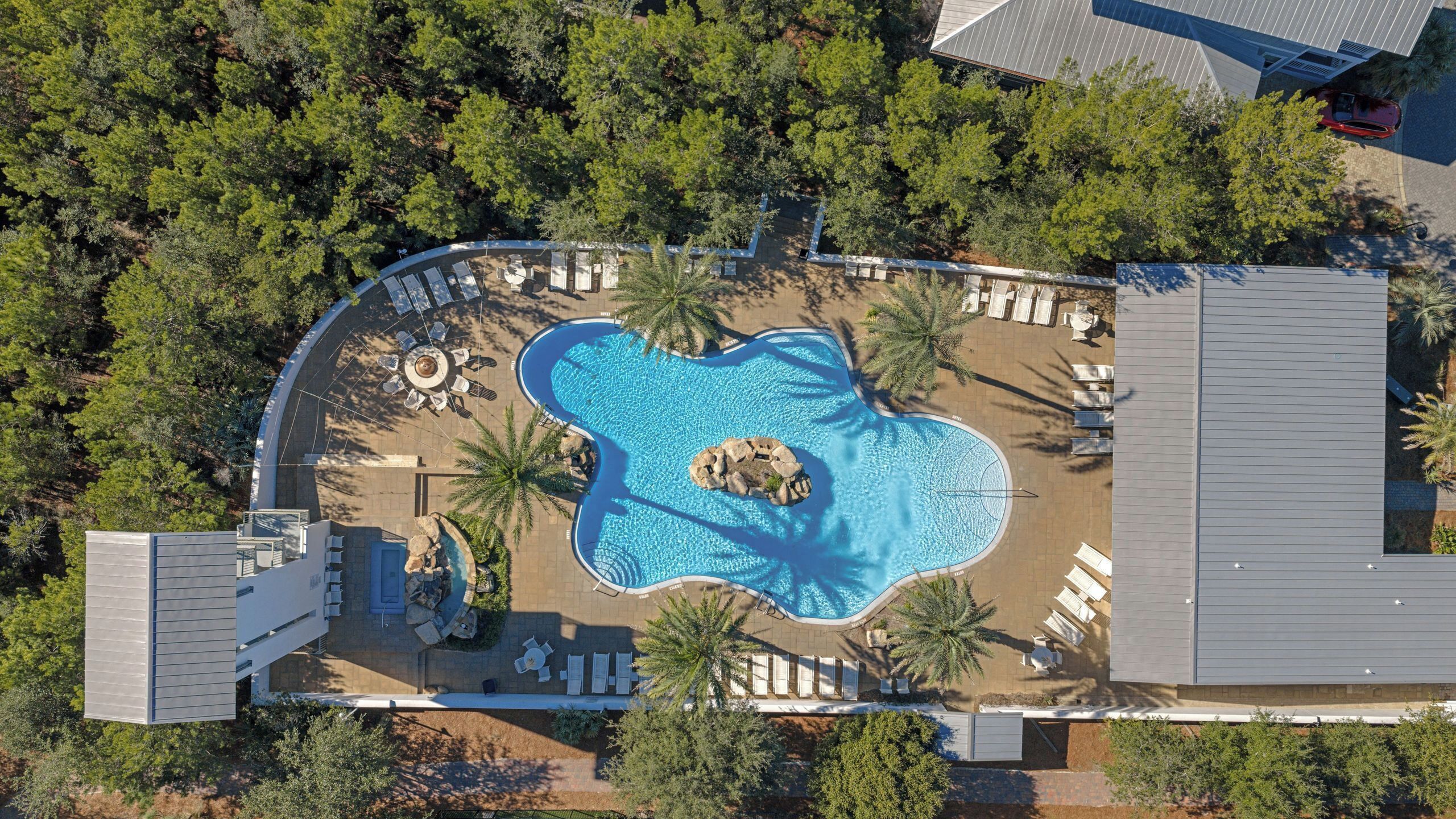 An aerial view of a resort with a central swimming pool, lounge areas, and surrounding trees.