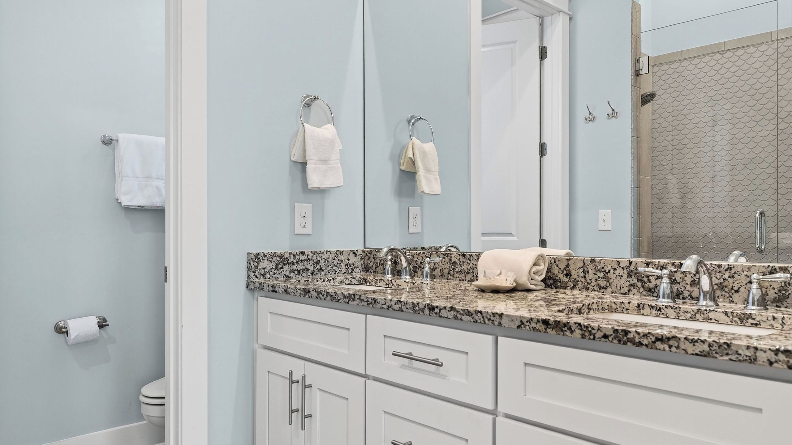 A bathroom with a granite countertop, two sinks, a mirror, a toilet, and a towel rack.