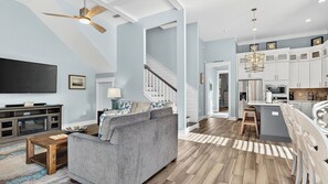 A spacious living area with a grey sofa, wooden coffee table, and a flat-screen TV. A staircase leads to a kitchen with white cabinets and a dining area. - 5min Walk to Dune Allen Beach Access Deck w/ Swing Charming 30A by Avantstay (Santa Rosa Beach)