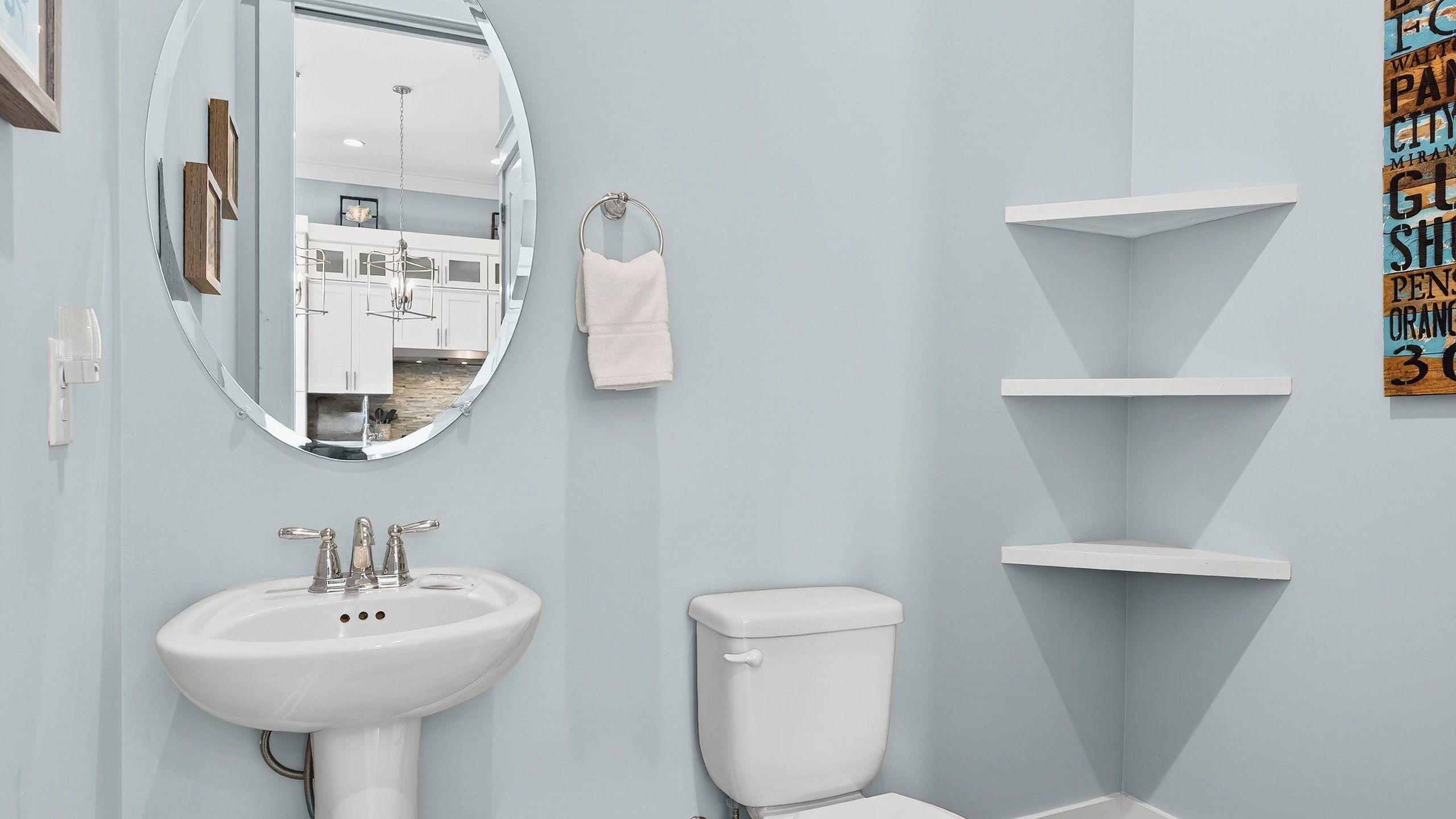 A bathroom with a white sink, toilet, and mirror. There are white shelves on the wall and a towel hanging on a ring.