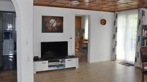 Un salon avec un plafond en bois, un téléviseur à écran plat, un tableau et une cuisine visible à travers une porte ouverte.