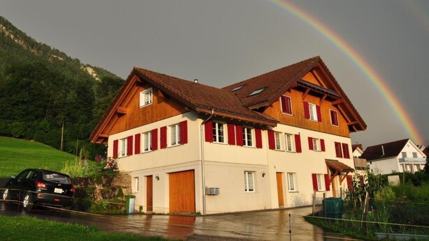 Un bâtiment à deux étages, doté d’un toit en pente et de volets rouges, entouré de verdure et avec une chaîne de montagnes en arrière-plan.