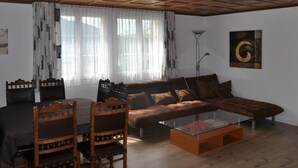 Un salon avec un plafond en bois, un canapé, deux fauteuils, une table basse en verre et un tableau accroché au mur.