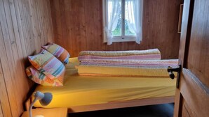 Une petite pièce chaleureuse aux murs en bois, avec un lit simple recouvert d’une literie colorée, un bureau avec une lampe et une fenêtre agrémentée de rideaux.