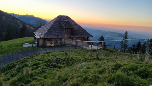 Un chalet de montagne rustique, doté d’un toit à forte pente, entouré de collines verdoyantes et sous un ciel dégagé.
