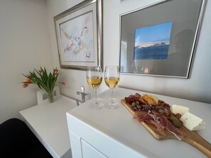 Ein Essbereich mit einem weißen Tisch, zwei Gläsern Weißwein, einer Holzplatte mit verschiedenen Käsesorten und Wurstwaren, einer Vase mit Tulpen und zwei gerahmten Bildern an der Wand.