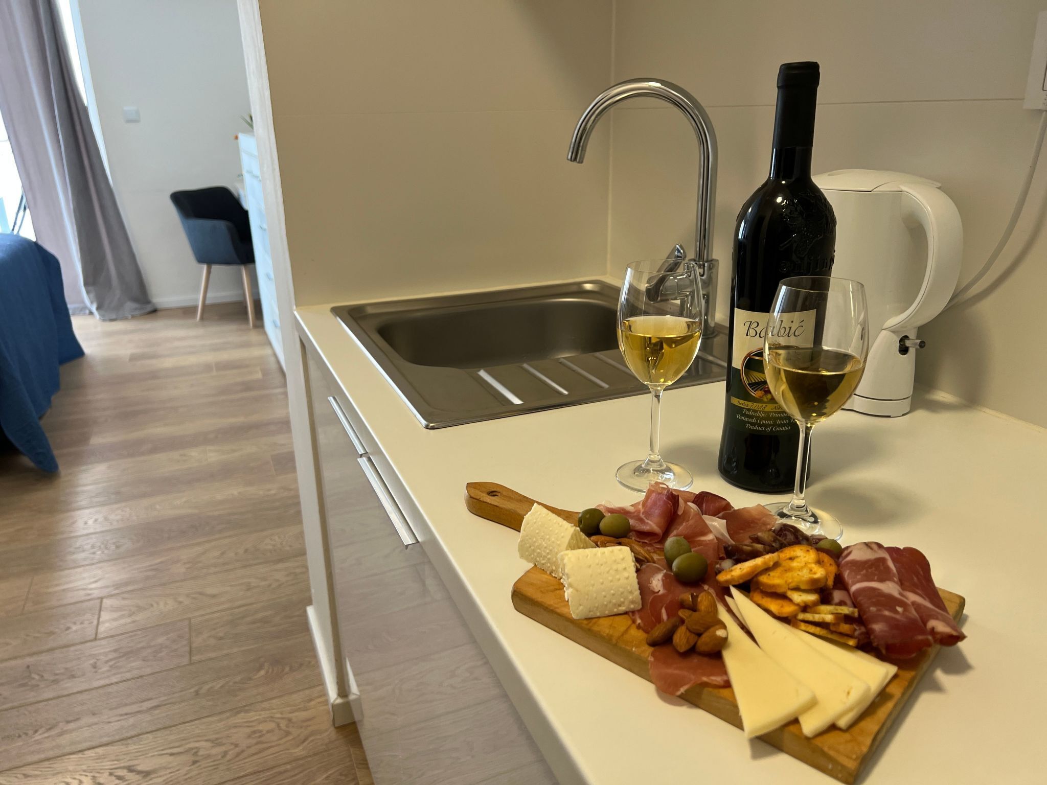 A kitchen with a white countertop, a stainless steel sink, a wine bottle, two glasses of white wine, and a charcuterie board with cheese, olives, almonds, and meats.
