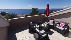 En terrasse med fletningsmøbler og en rød parasol, med udsigt over kysten.