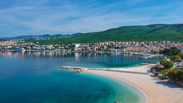 En kystby med en sandstrand, klart blåt vand og et tætbebygget område med bygninger og bakker i baggrunden.