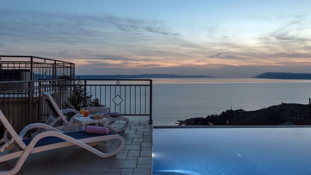 Eine Terrasse mit Lounge-Möbeln, einem Tisch mit Getränken und Blick auf das Meer bei Sonnenuntergang.