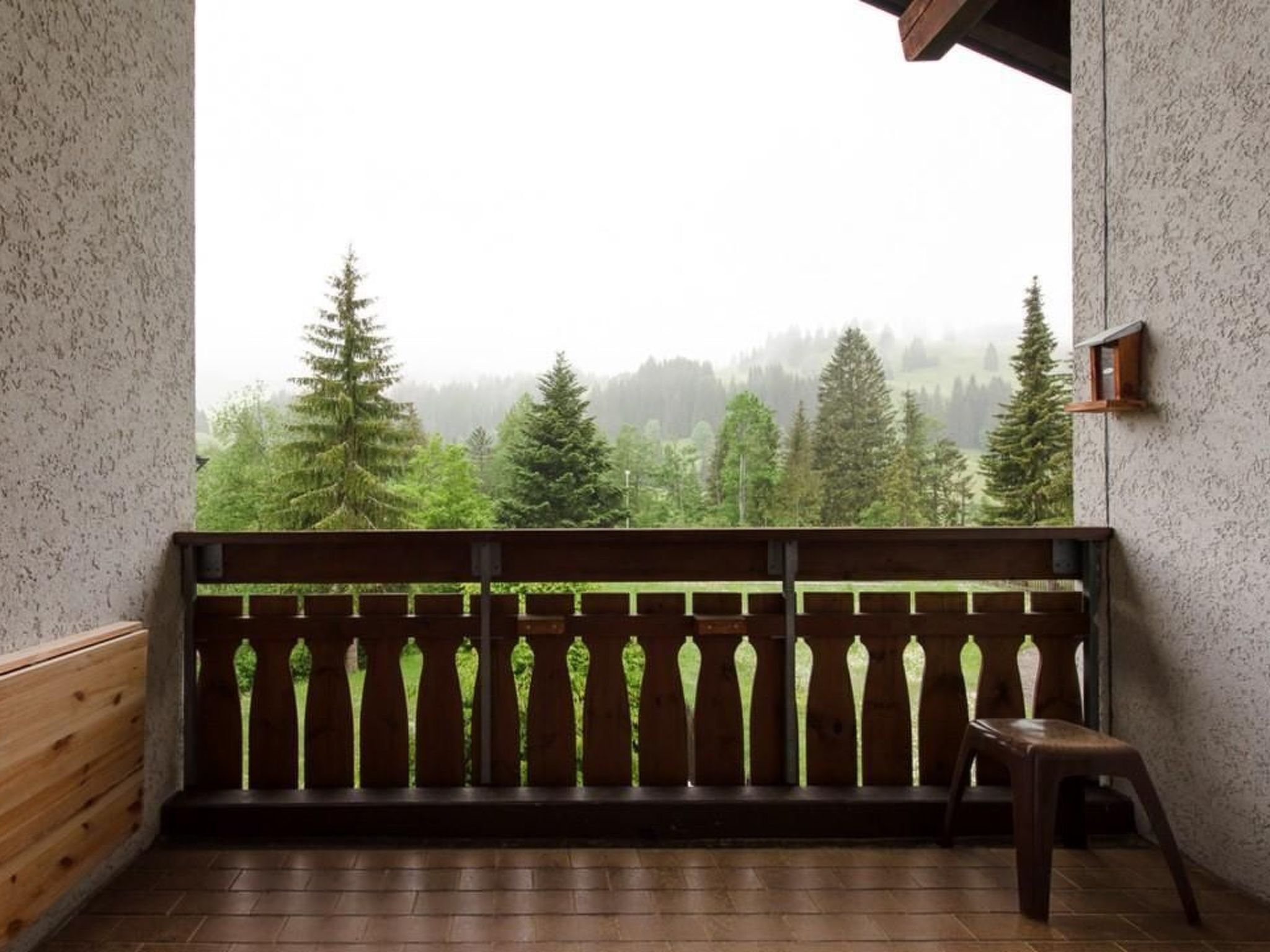 A balcony with a wooden railing, a small stool, and a view of a forested landscape through a misty window.