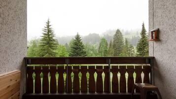 A balcony with a wooden railing, a small stool, and a view of a forested landscape through a misty window.