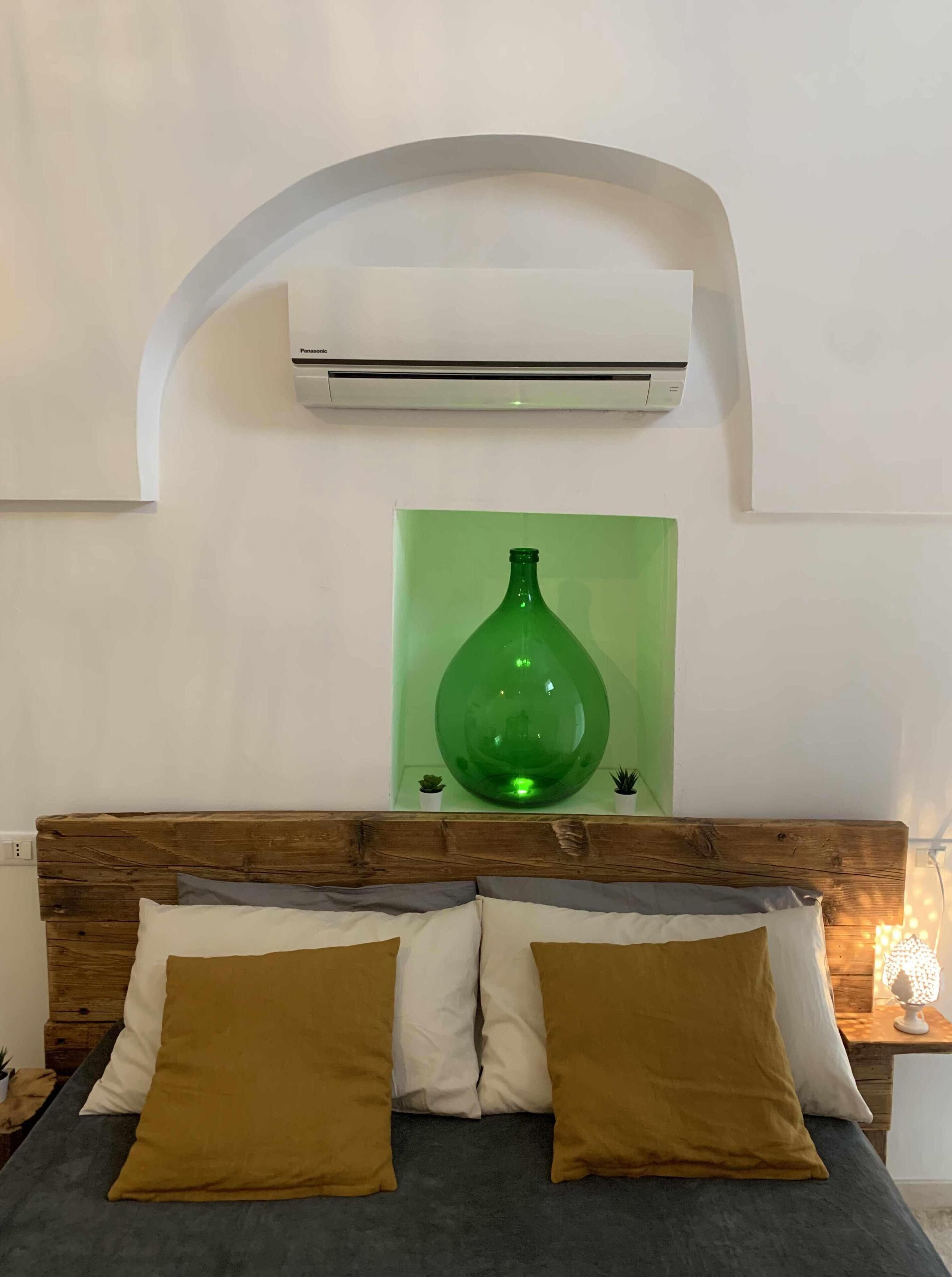 Un dormitorio con cabecera de madera, una cama con dos almohadas, un jarrón verde en un estante y un equipo de aire acondicionado en la pared.