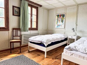 Ein Schlafzimmer mit zwei Betten, einem Sessel, einem Heizkörper, einem Fenster mit Vorhängen und einem Bild an der Wand.