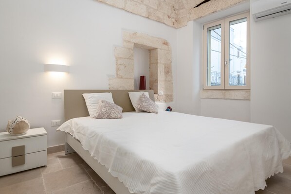 A bedroom with a bed, nightstand, lamp, and a stone archway. - Le Volte, Le Piccole Case Bianche (Ostuni)