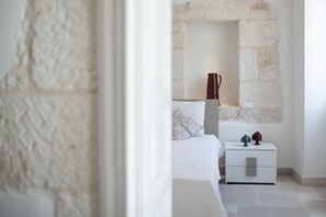 A bedroom with a stone wall, a bed with white bedding, a bedside table with a lamp, and a nightstand with decorative items. - Le Volte, Le Piccole Case Bianche (Ostuni)