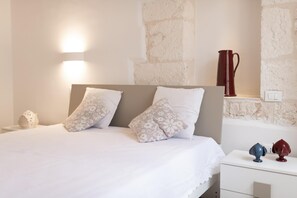 A bedroom with a bed, pillows, a nightstand, a red pitcher, and a wall with a textured pattern. - Le Volte, Le Piccole Case Bianche (Ostuni)