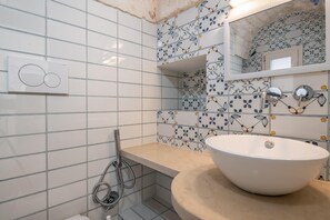 A bathroom with a white tiled wall, a beige countertop, a white sink, a mirror, and a shower. - Le Volte, Le Piccole Case Bianche (Ostuni)