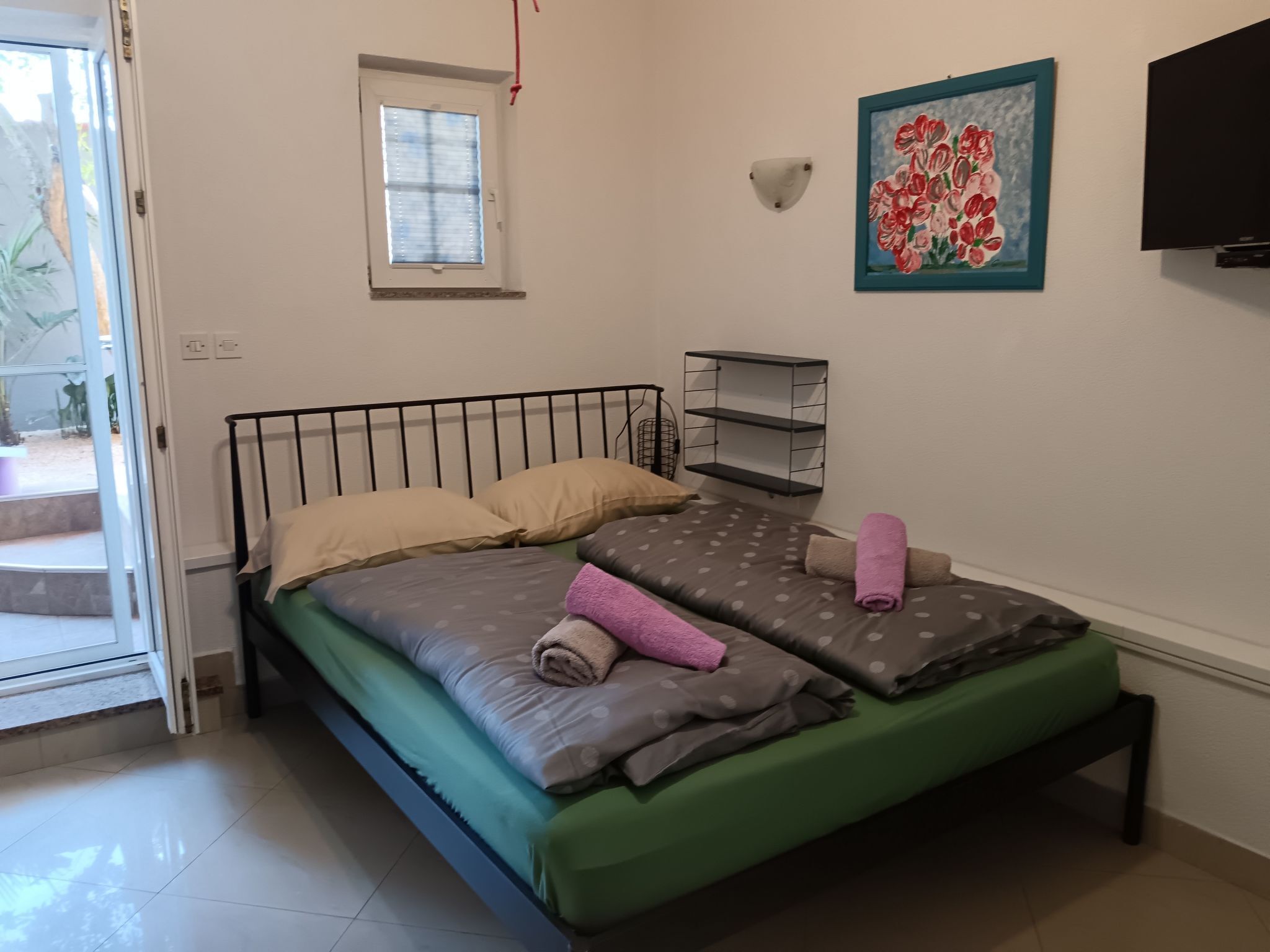 A bedroom with a bed, pillows, a green mattress, a window, a wall-mounted shelf, and a painting.