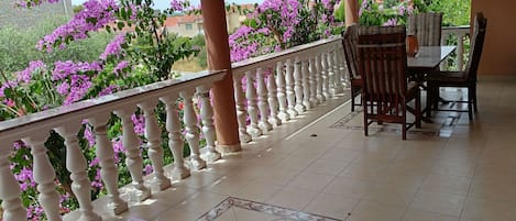 A balcony with a white balustrade, a dining table with chairs, and purple flowers in the background.