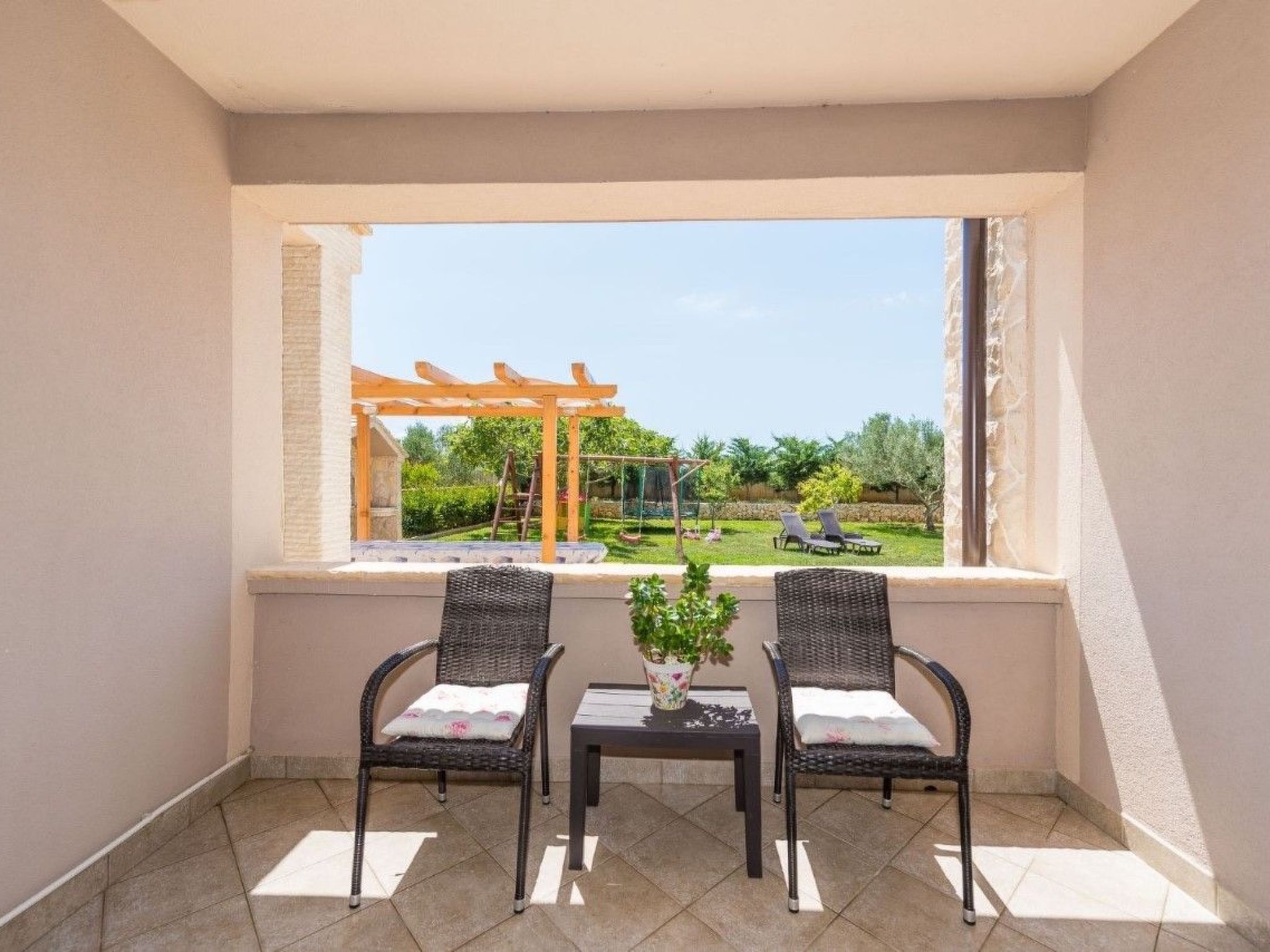 A balcony with two wicker chairs and a small table, a wooden pergola, and a view of a garden.