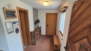 Ein Flur mit einer Holztür, einer Sitzbank und einem gefliesten Boden. An der Wand befinden sich Schilder und links eine Treppe.
