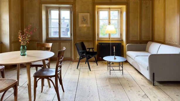 Una habitació amb una taula rodona de fusta, dues cadires, un sofà blanc i una taula petita rodona amb una làmpada. Hi ha dues finestres amb cortines, una de les quals té un radiador a sota.