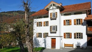 Edifici blanc amb persianes de fusta i teulada vermella, envoltat de vegetació i turons.