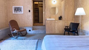 Una habitació d'hotel moderna amb interior de fusta, un llit, una cadira d'oscil·lació, un escriptori amb cadira, un bany amb rentapans i mirall, i un quadre amb marc penjat a la paret.