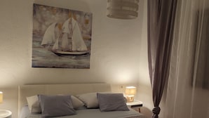 A bedroom with a bed, pillows, a nightstand, a lamp, a painting of sailboats, and a window with curtains.